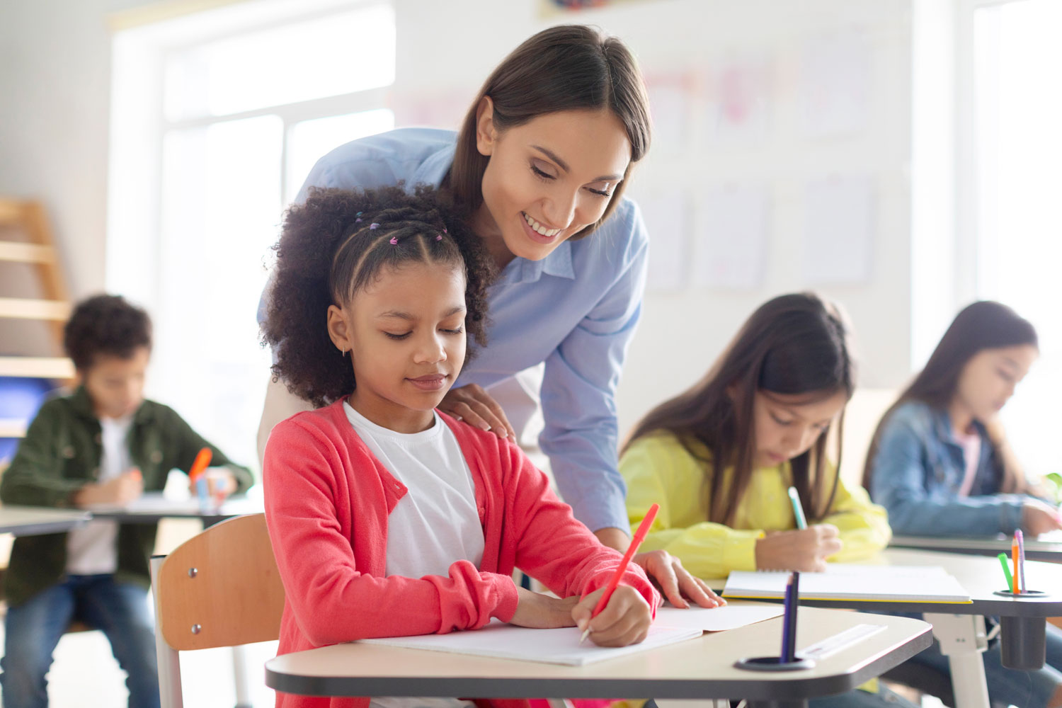 Cheerful female teacher helping girl with homework in classroom, black schoolgirl writing in notebook, sitting at desk with classmates on background